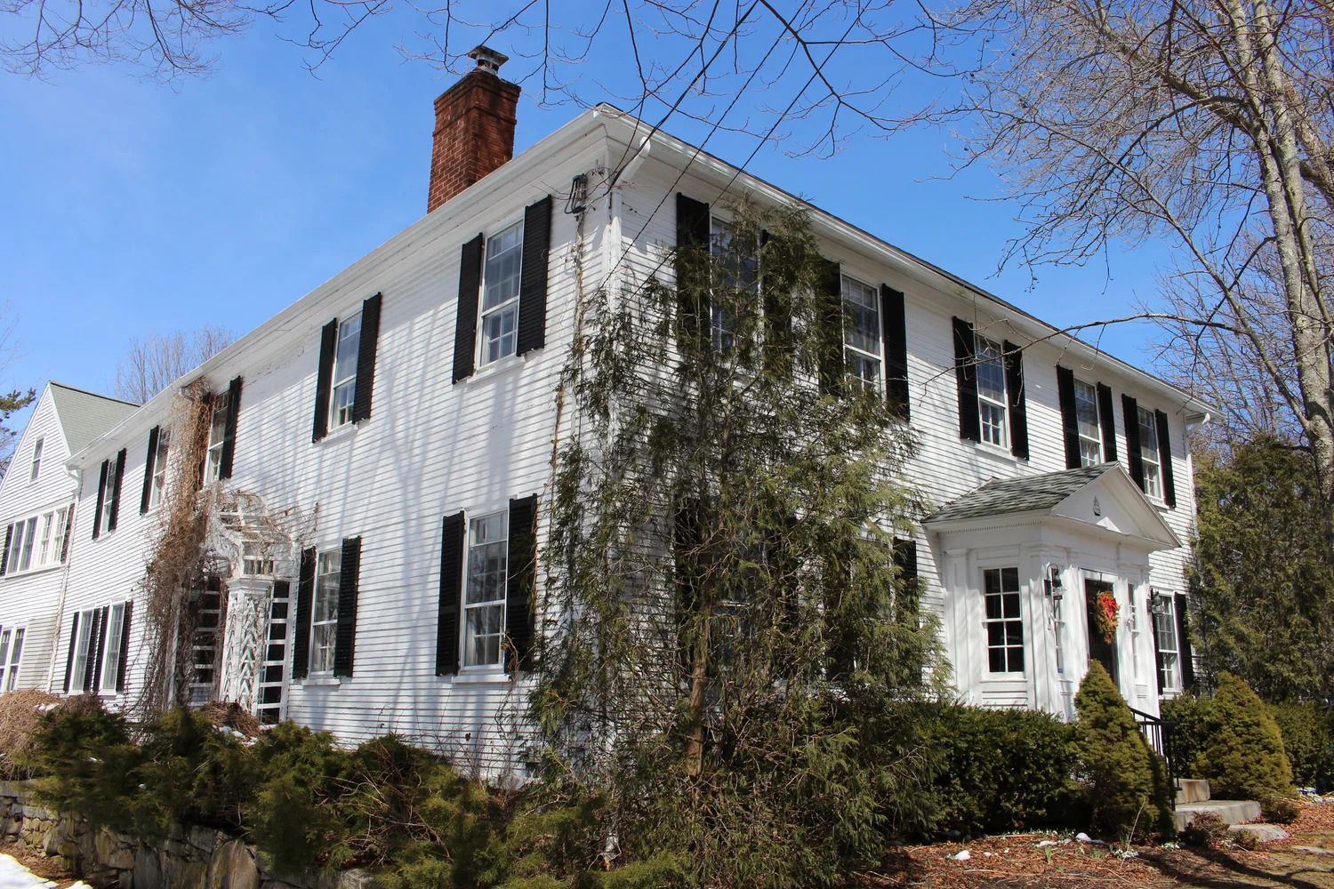 White two-story house with black shutters, brick chimney, and shrubs. Bright sunny day.