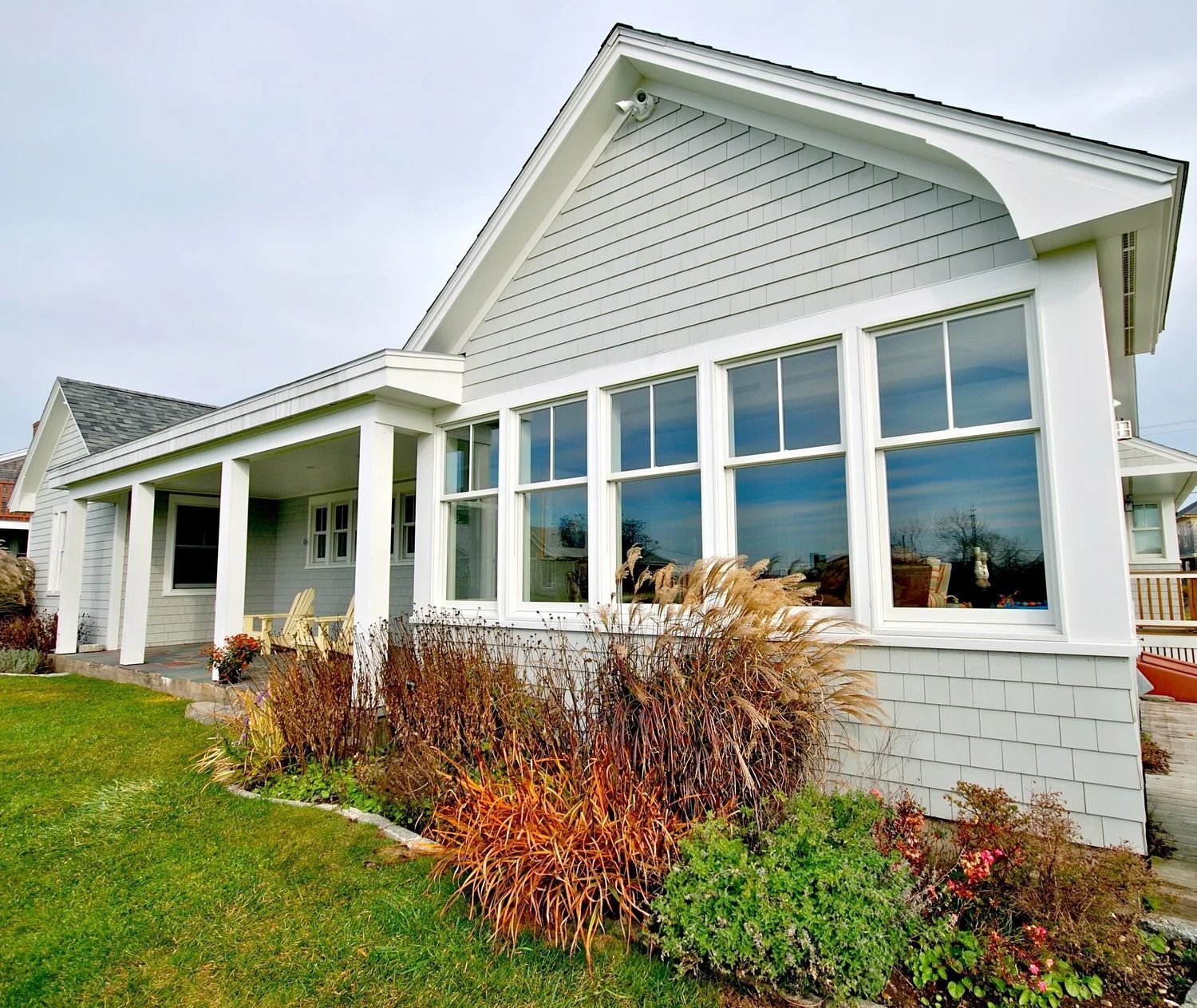 Light gray coastal home with large windows and porch, surrounded by green grass and colorful bushes.