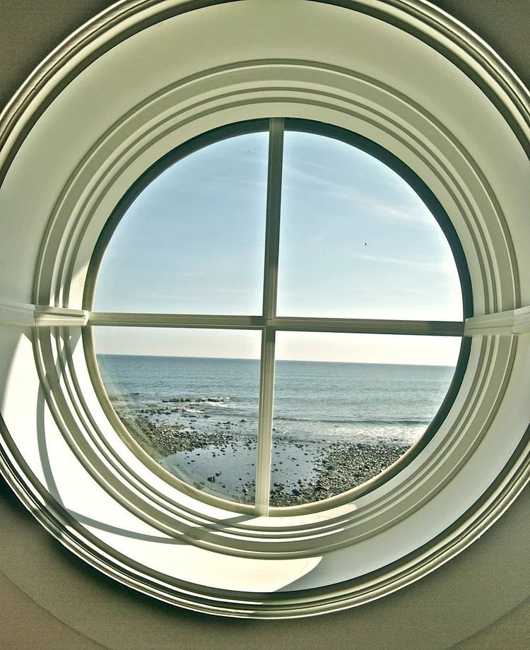 Round window framing a view of the ocean and beach on a sunny day.