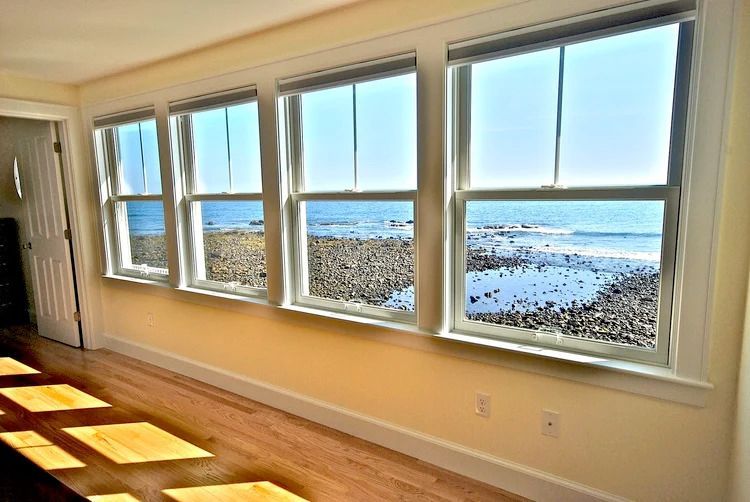 Coastal room with four large windows overlooking a rocky beach and ocean; bright sunlight.