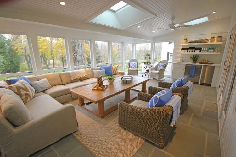 Sunroom with a beige sectional, wicker chairs, wooden coffee table, and large windows overlooking trees.