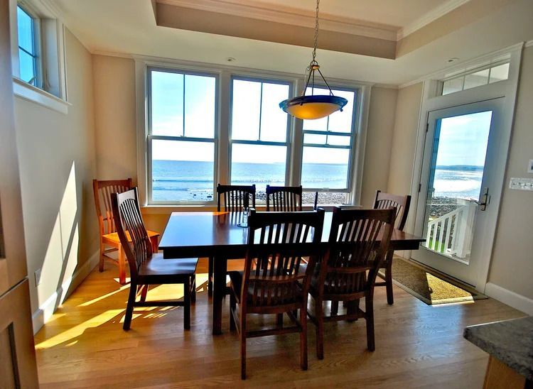 Dining room with large windows overlooking a beach and ocean. A dark wooden table and chairs sit in the room.
