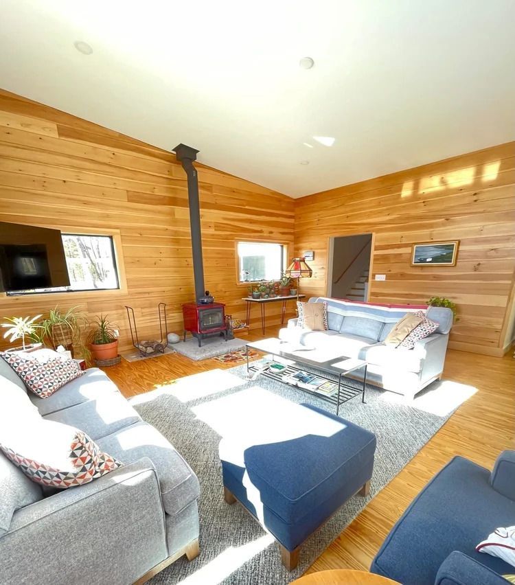 Cozy living room with wood-paneled walls, gray sofas, blue ottoman, fireplace, and natural light.