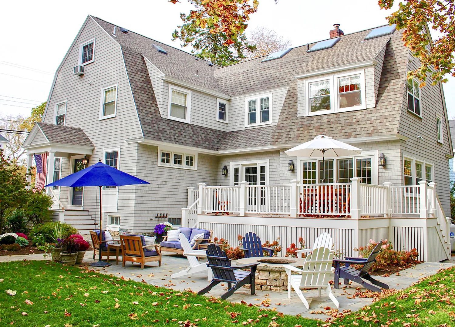 Backyard patio of a large two-story house with a wooden deck, adirondack chairs, and umbrellas.