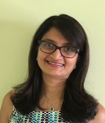 戴著眼鏡的女人面帶微笑,黑髮,白皮膚,穿著有圖案的上衣,靠著一面綠色的牆壁站著。