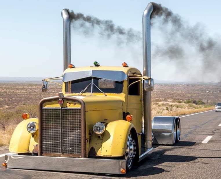 A yellow semi truck with smoke coming out of its exhaust pipes is driving down a road.