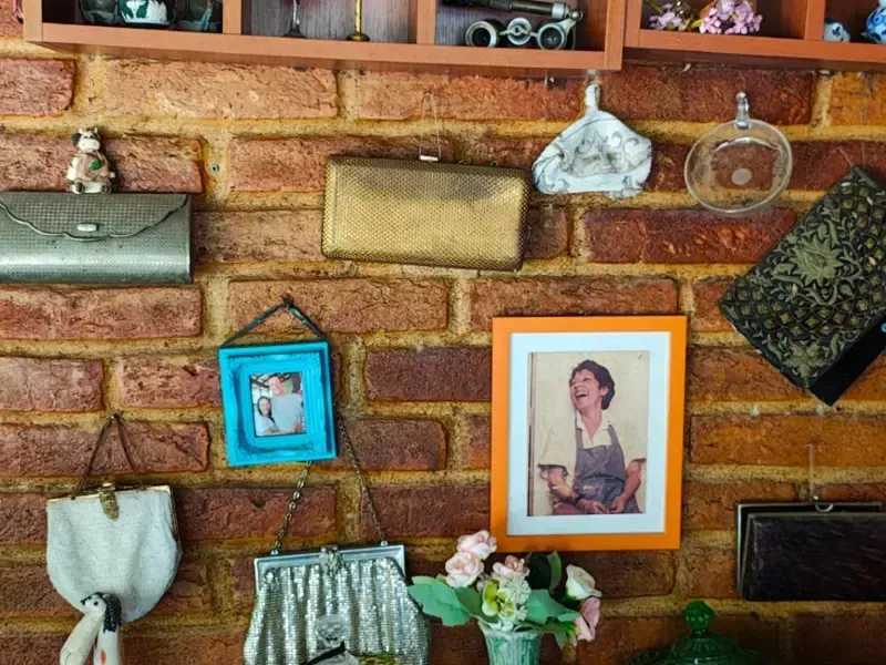 Parede de tijolos com bolsas penduradas, fotos emolduradas, um casaco e um pequeno vaso de flores.