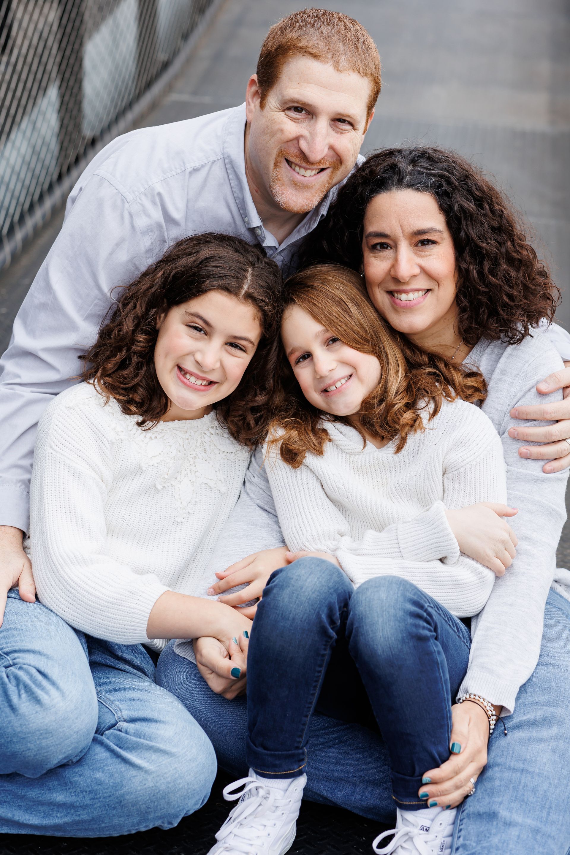 Family of four smiling, posing outdoors. Two girls in white sweaters, parents embrace, smiling.