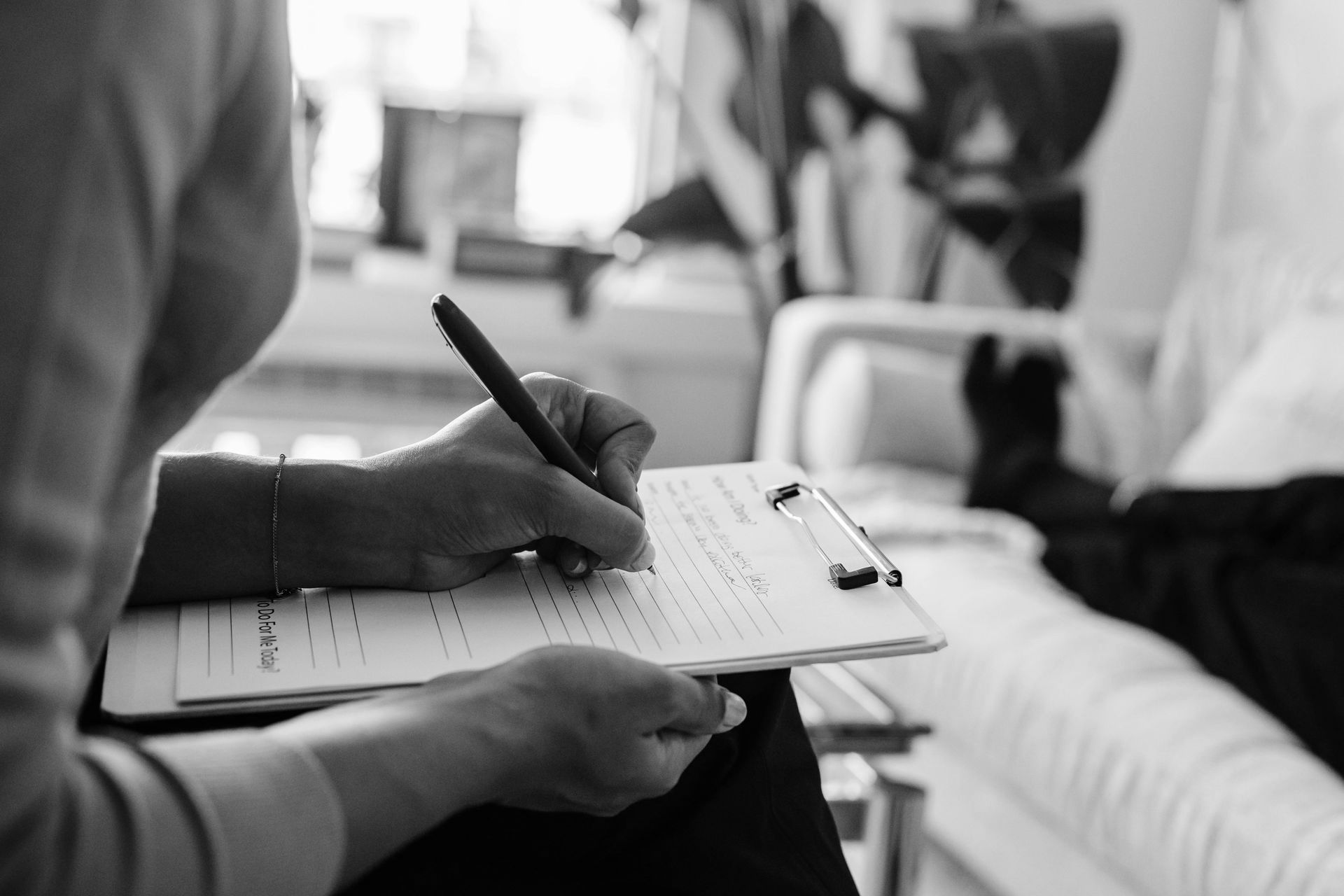Person taking notes on a clipboard while another lies in a hospital bed. Black and white.