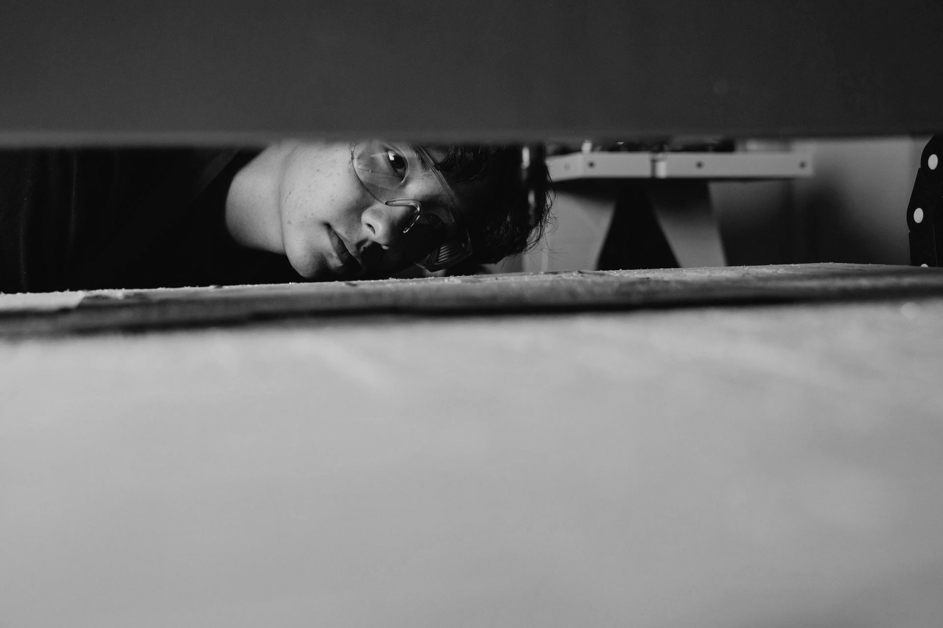 A person peering through a gap in a dark room, head resting on a surface, in black and white.