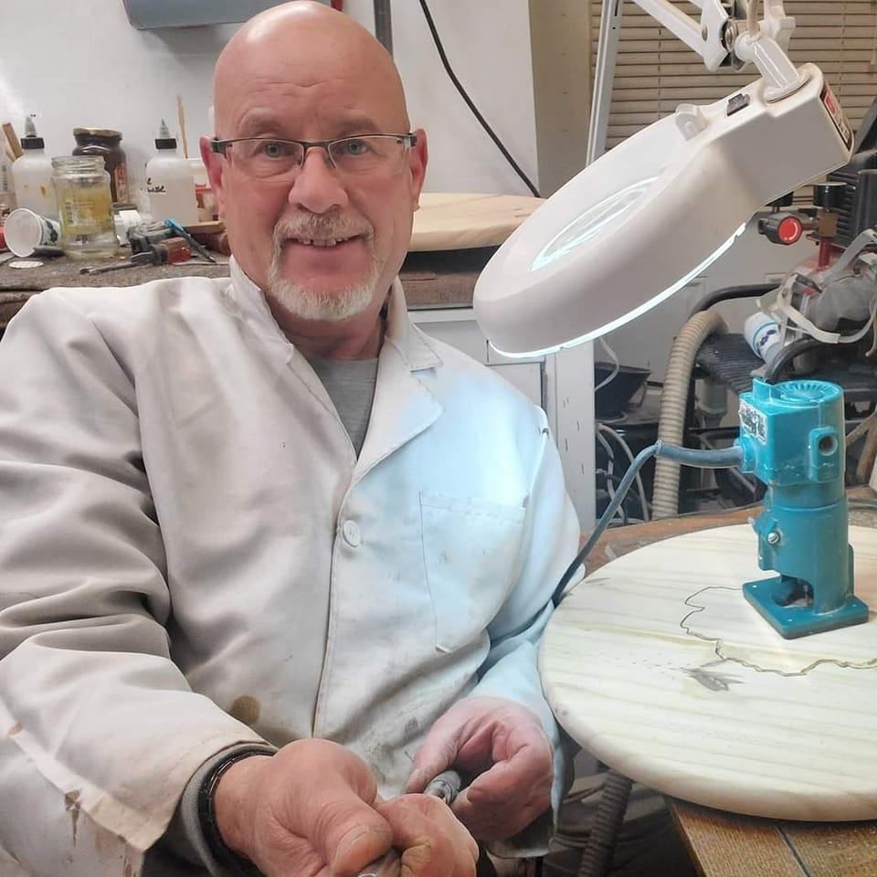 Man with glasses and lab coat smiles while working on wood carving, lit by lamp.