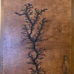 Black fractal-like Lichtenberg figure burned into wood. Brown tones and vertical design.