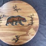 Wooden round with bear and trees carving, brown and black details.