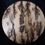A round wooden panel with burnt fractal patterns resembling trees in black against the light wood grain.