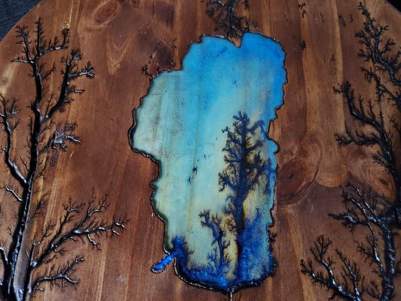 Wooden art with blue lake, burned trees on brown wood.