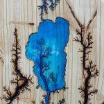 Blue painted lake with black, burnt-wood trees on a light wood background.
