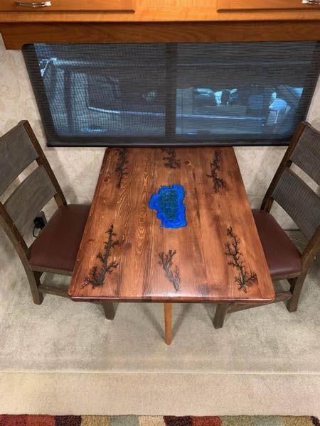 Wooden table and two chairs in RV interior. Table has blue epoxy and etched design. Dark brown chairs.