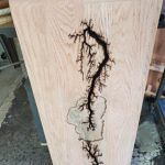 Wooden slab with a dark, fractal-like Lichtenberg figure burned into its surface, resembling lightning.