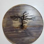 Circular wood piece with a burned tree design. Gray and brown tones.