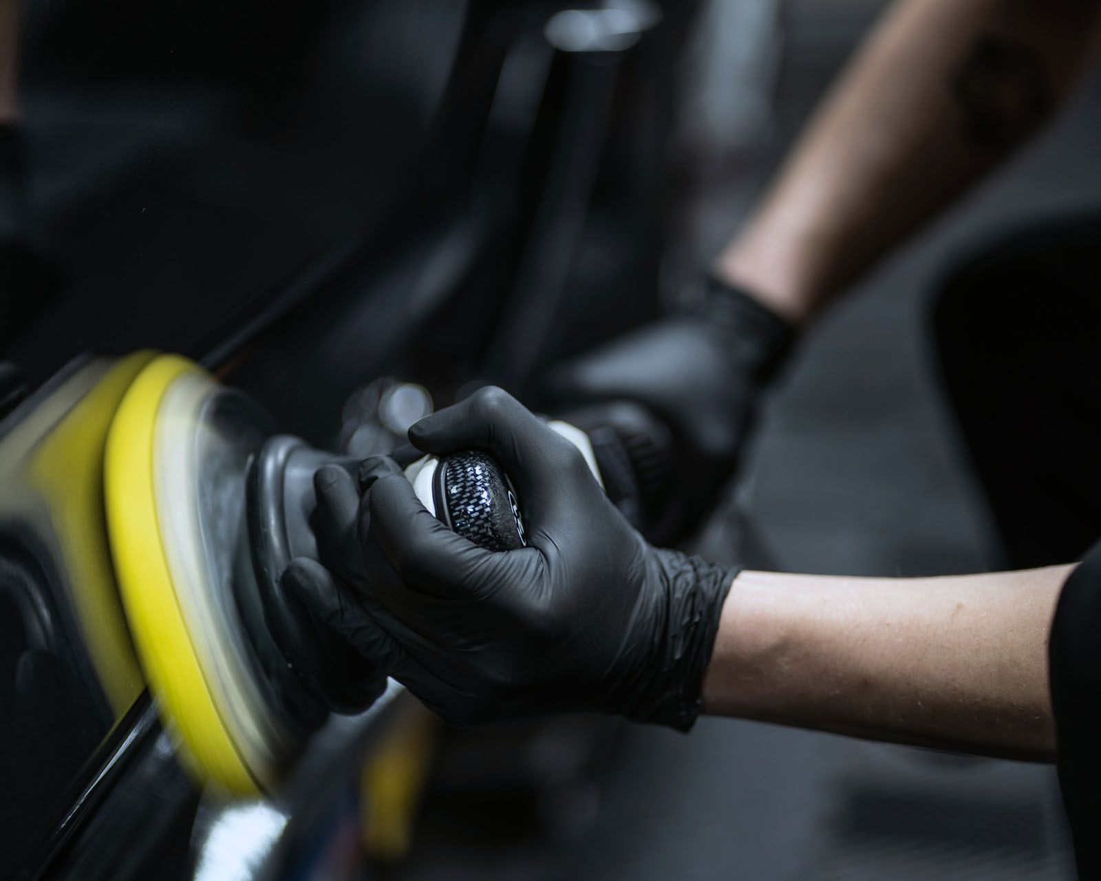 A person wearing black gloves is polishing a car with a yellow polisher.