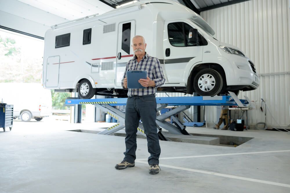 A Man Is Standing In Front Of A Rv On A Lift In A Garage — Affordable Caravan Repairs in Bundaberg, QLD