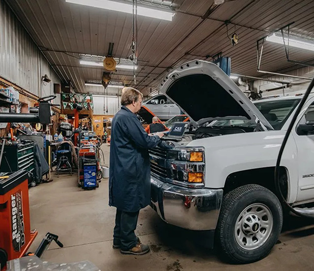 Mechanic in blue coat, using laptop to diagnose a white truck's engine in a garage. | Auto Source Of West Michigan