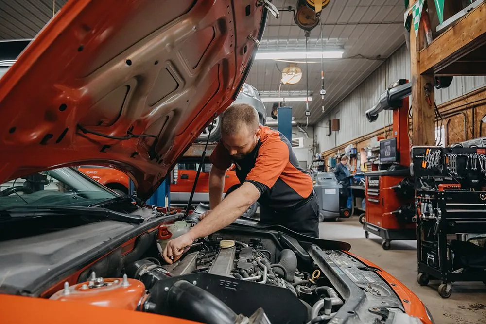 Mechanic working on an orange car engine in a garage. | Auto Source Of West Michigan