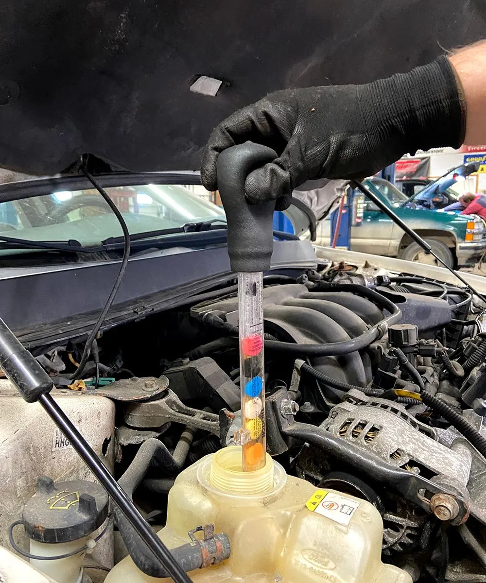 Gloved hand testing car coolant with a hydrometer in an engine compartment, displaying color-coded levels. | Auto Source Of West Michigan