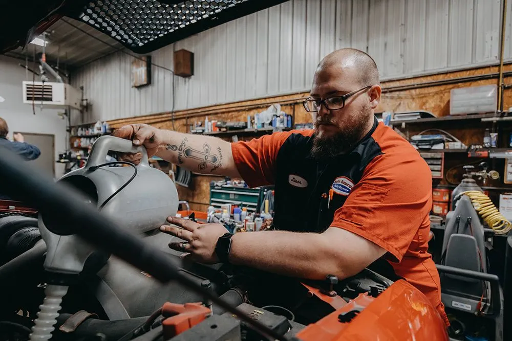 Mechanic in red shirt works on a car engine inside a cluttered garage. | Auto Source Of West Michigan