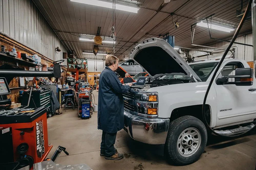 Mechanic in a blue coat working on a white truck with the hood open inside a garage. | Auto Source Of West Michigan