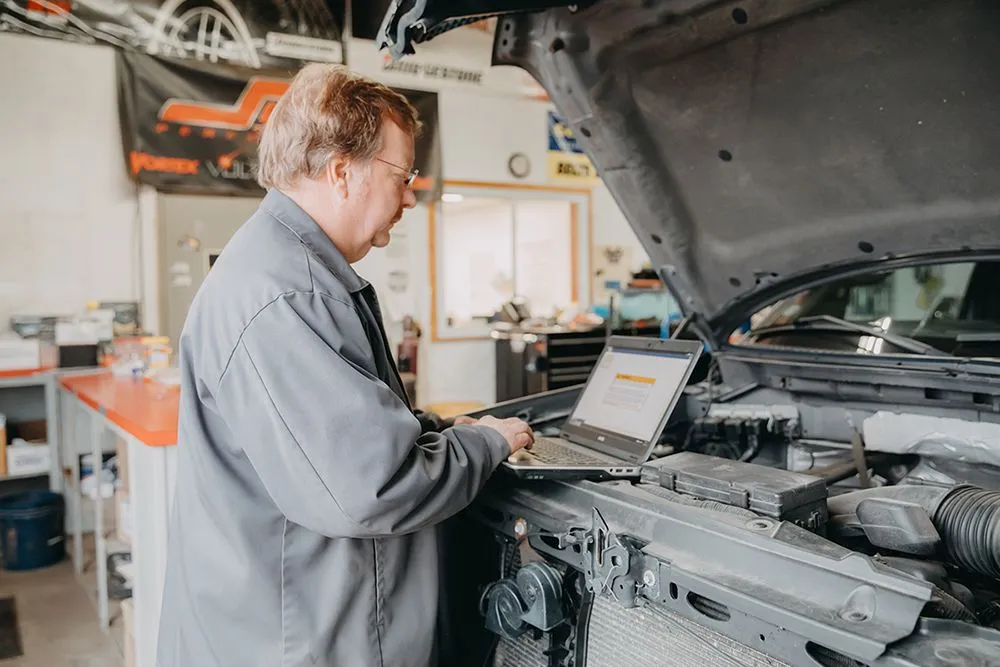 Mechanic in gray overalls uses a laptop to diagnose a car engine in a garage. | Auto Source Of West Michigan