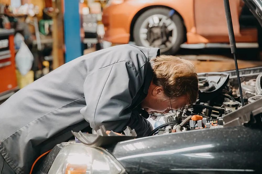 Mechanic in grey uniform working on a car engine in a garage with an orange car in the background. | Auto Source Of West Michigan