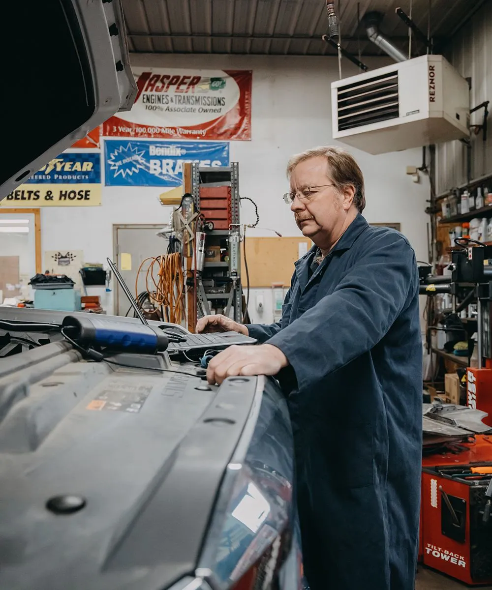 Mechanic in blue coveralls working on a car engine in a garage. | Auto Source Of West Michigan
