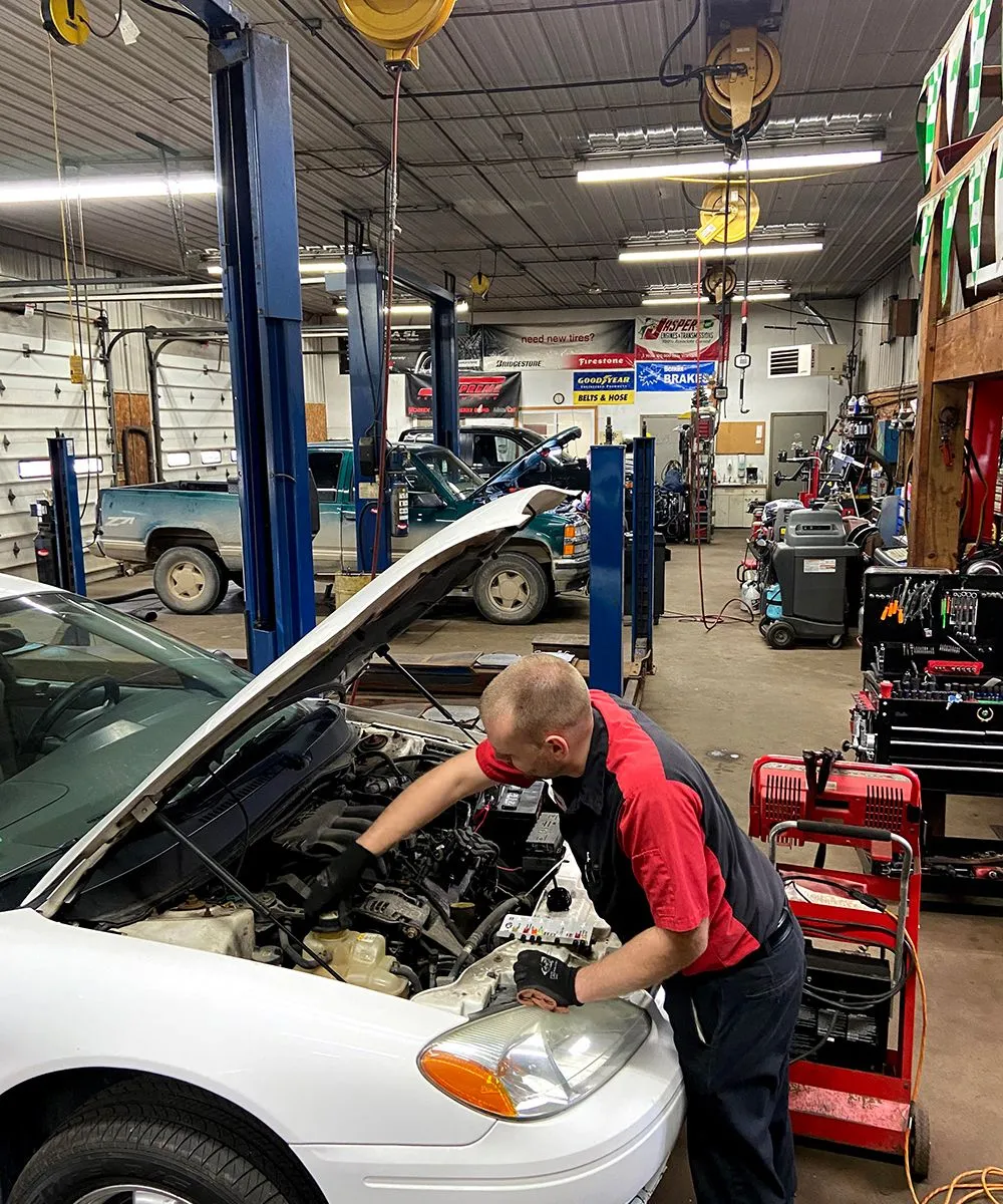 Mechanic working on a white car in a garage, wearing red and black uniform, with tools. | Auto Source Of West Michigan