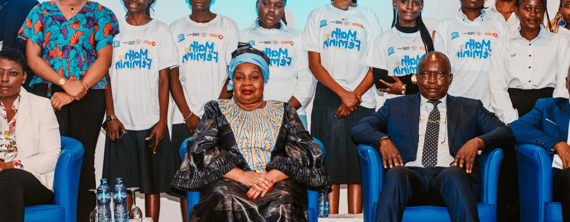Des personnes sont assises sur des chaises bleues devant un groupe. Une femme voilée sourit. Un homme en costume regarde droit devant lui.