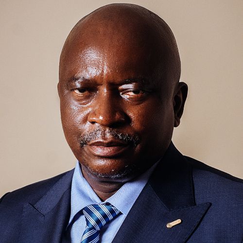 Homme en costume bleu, bras croisés, souriant ; fond en panneau de bois.