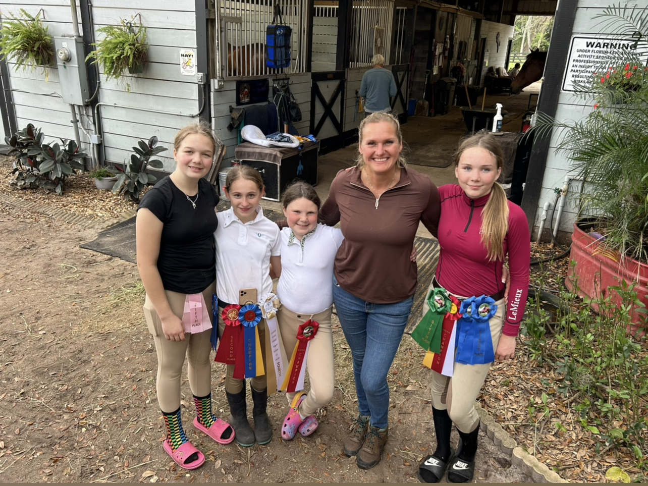 A group of young girls are posing for a picture with a woman in front of a horse stable.