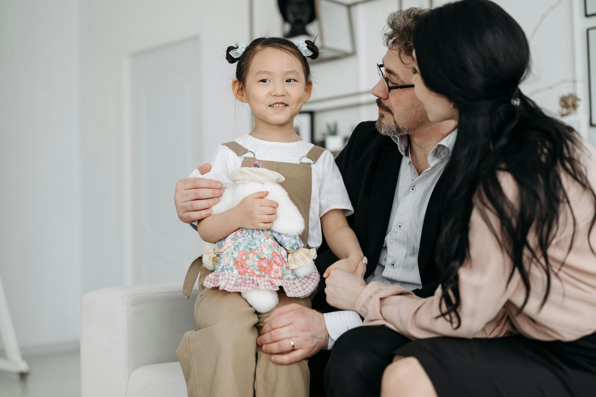 Jong meisje met staartjes houdt een knuffeldier vast en zit met een man en een vrouw op een bank.
