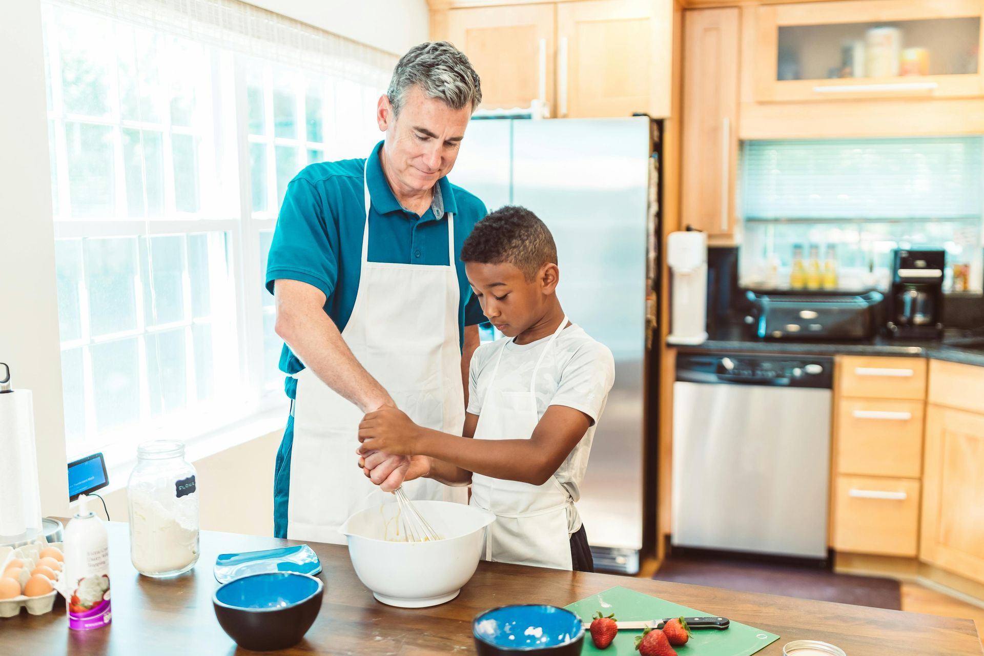 Vader en zoon bakken samen in de keuken; de jongen roert het beslag en de vader kijkt toe.
