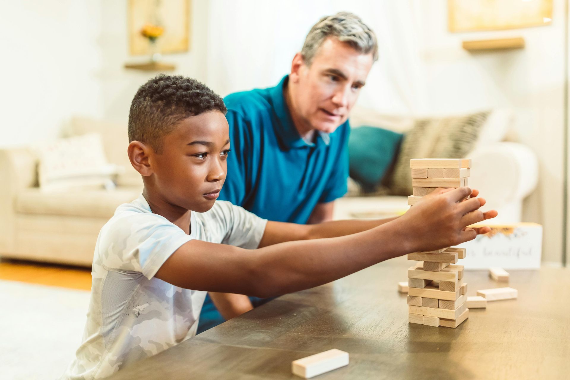 Een jongen en een oudere man spelen Jenga aan een tafel binnenshuis. De jongen concentreert zich terwijl hij een blokje trekt, de man kijkt toe.
