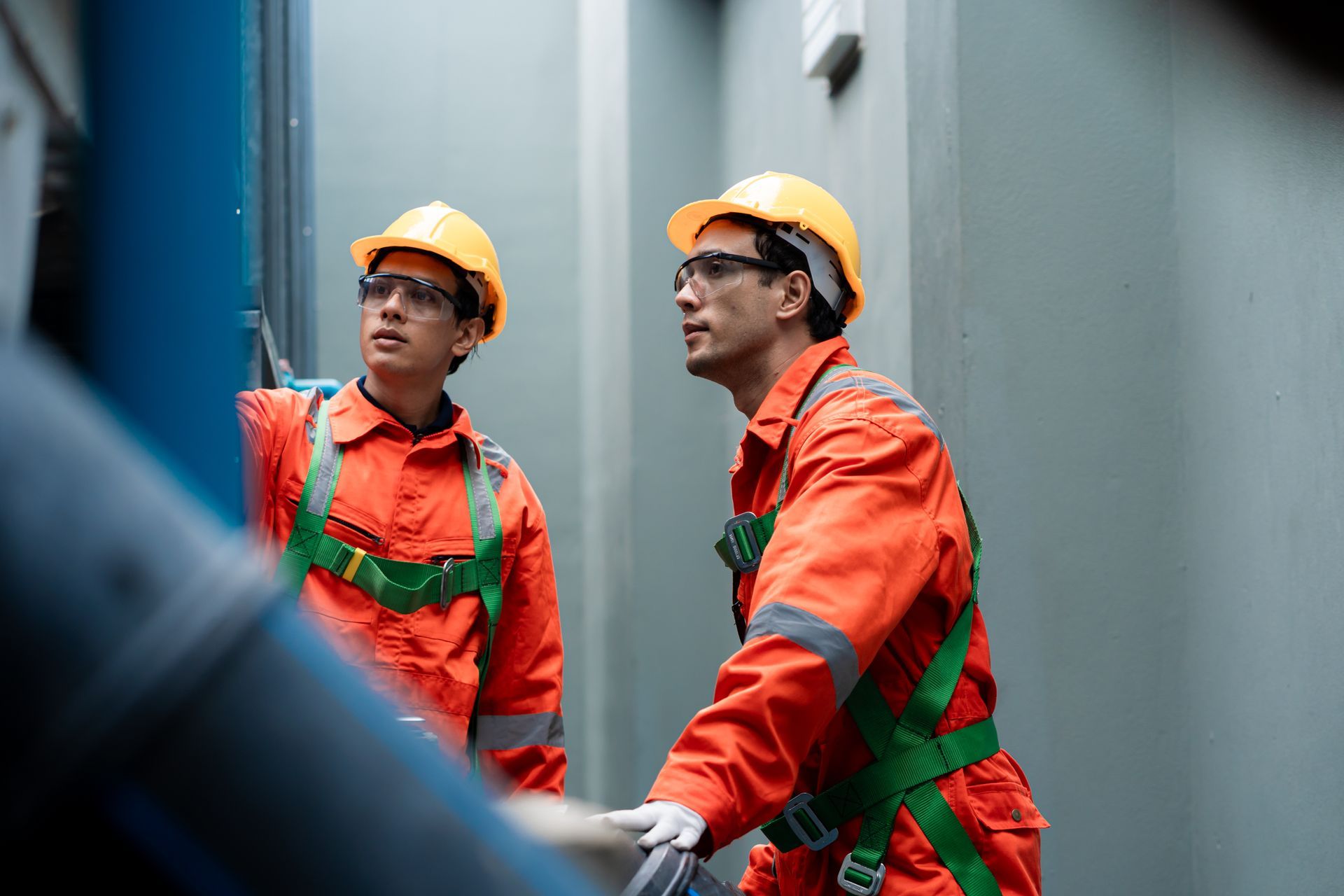 Two workers in orange safety gear and hard hats inspecting equipment in a narrow industrial area
