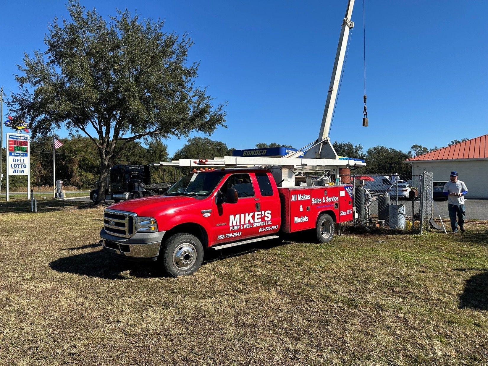 Red Mike’s Pump & Well Service truck with a crane rig parked on a grassy lot next to a Sunoco gas station, performing well or pump service work under a clear blue sky.