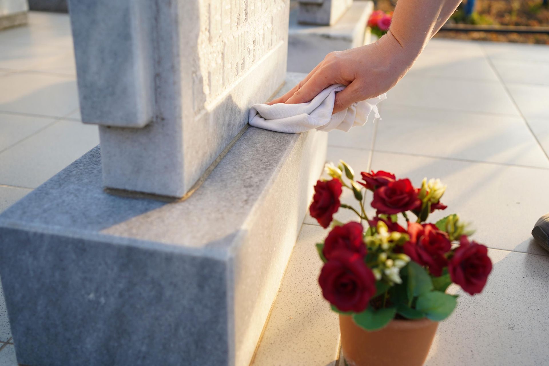Hand cleaning a marble headstone with a cloth, next to a pot of red roses.