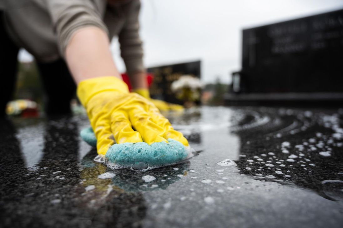Person wearing yellow gloves cleans a black granite headstone with a blue sponge in a cemetery.