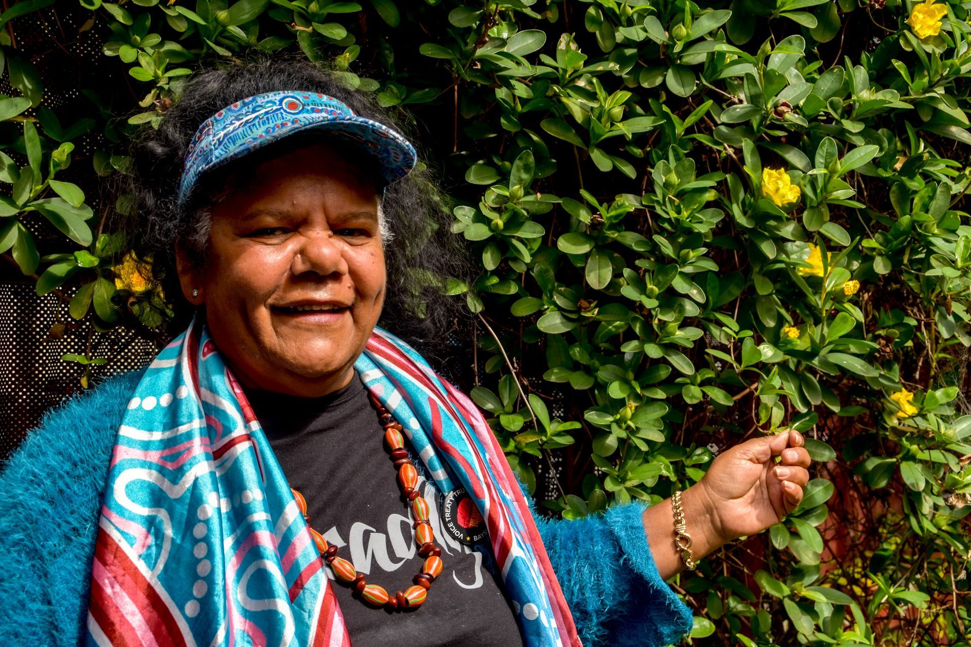 A woman wearing a hat and scarf is standing in front of a bush with yellow flowers.