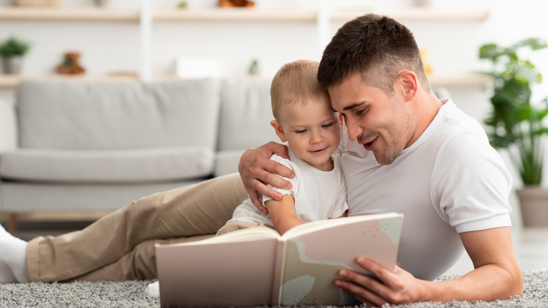 Father and child reading a book together on the floor. White shirt, neutral setting, smiling faces.