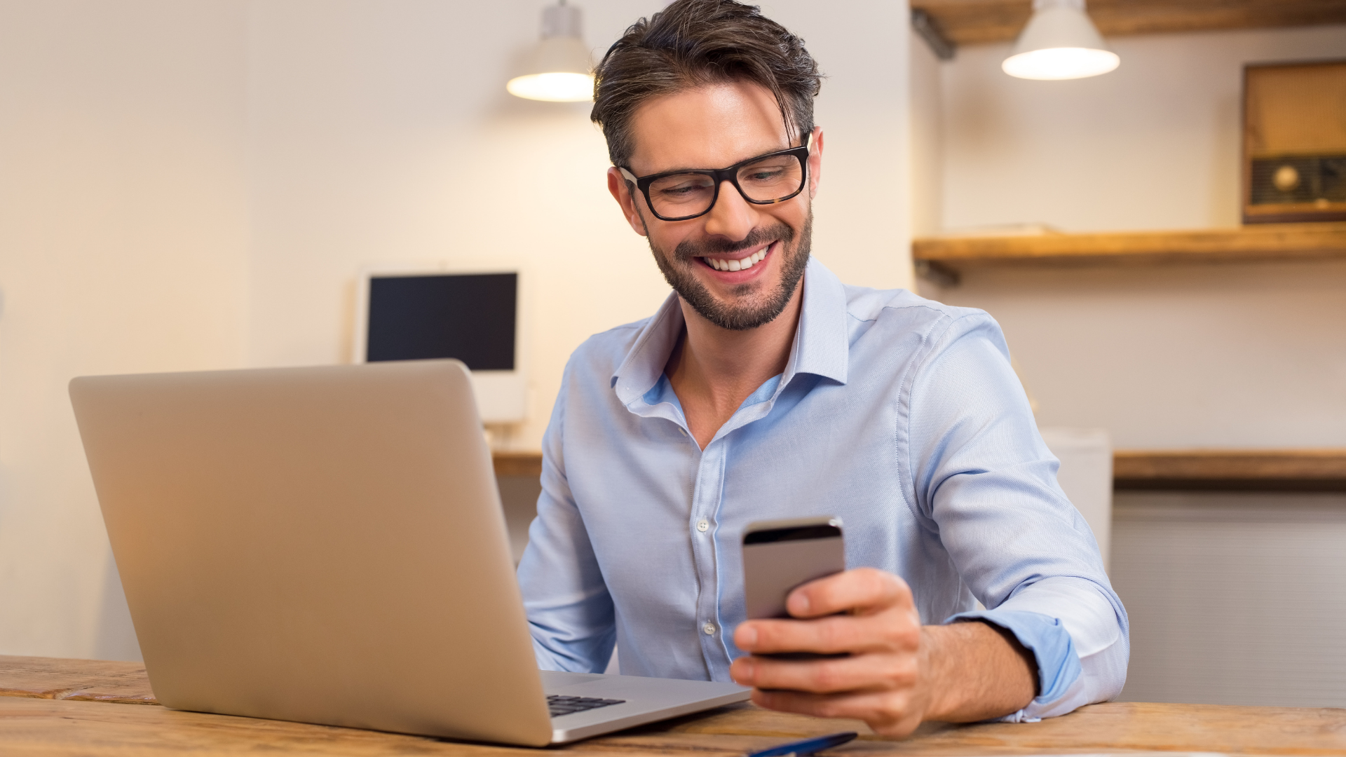 Man with glasses smiles while using a smartphone and laptop at a desk.