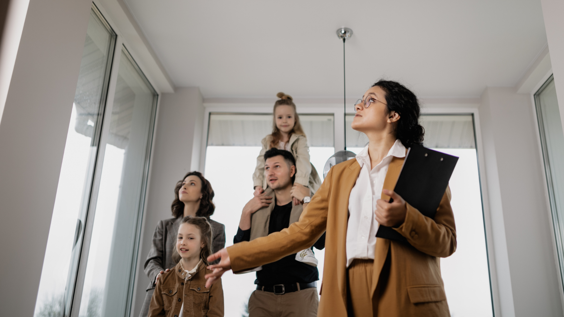 Real estate agent showing a family a home's interior. Agent gestures towards windows; everyone looks on.