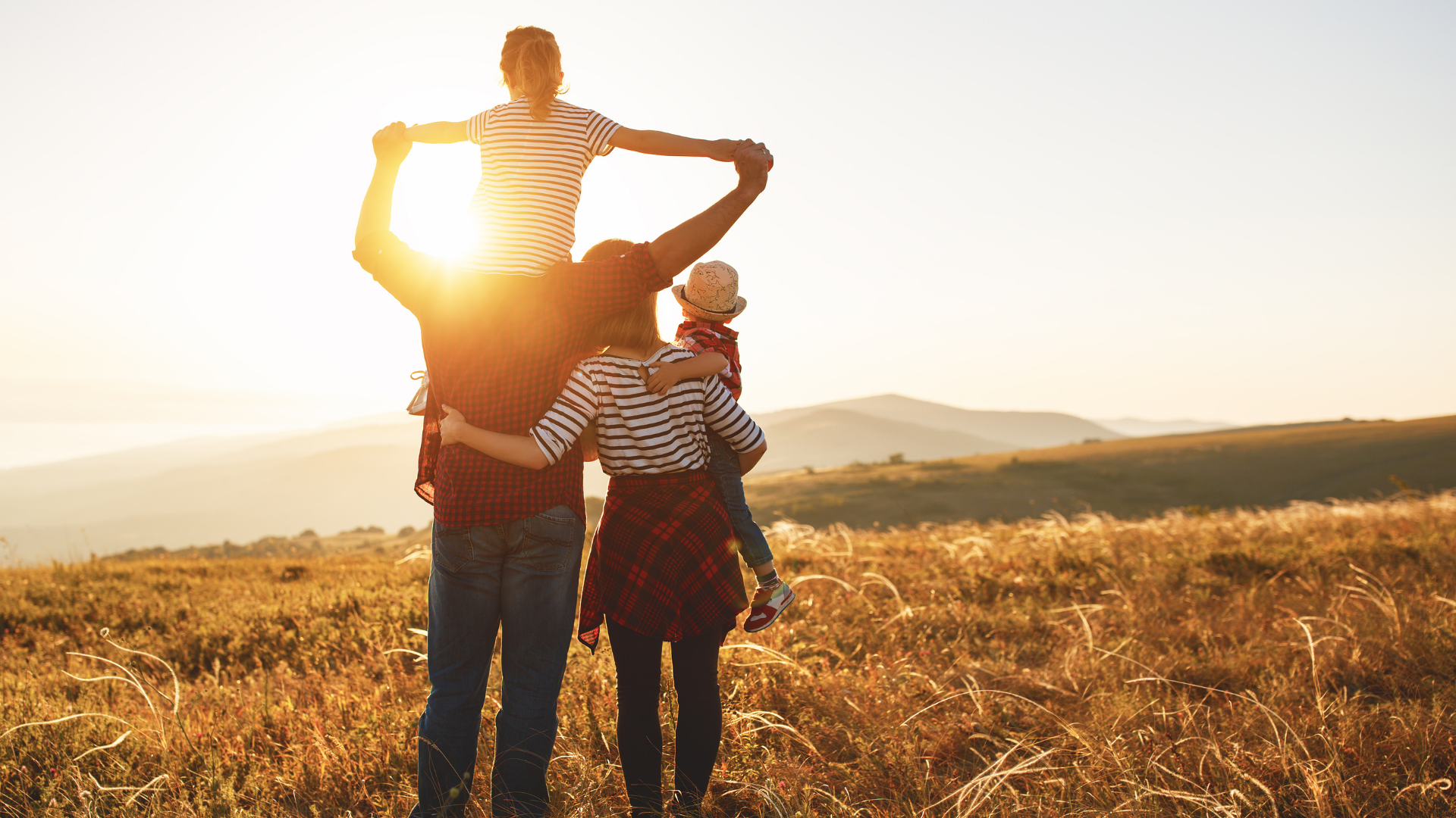 Family of four walking in a field at sunset; father carries a child on his shoulders, mother embraces another.