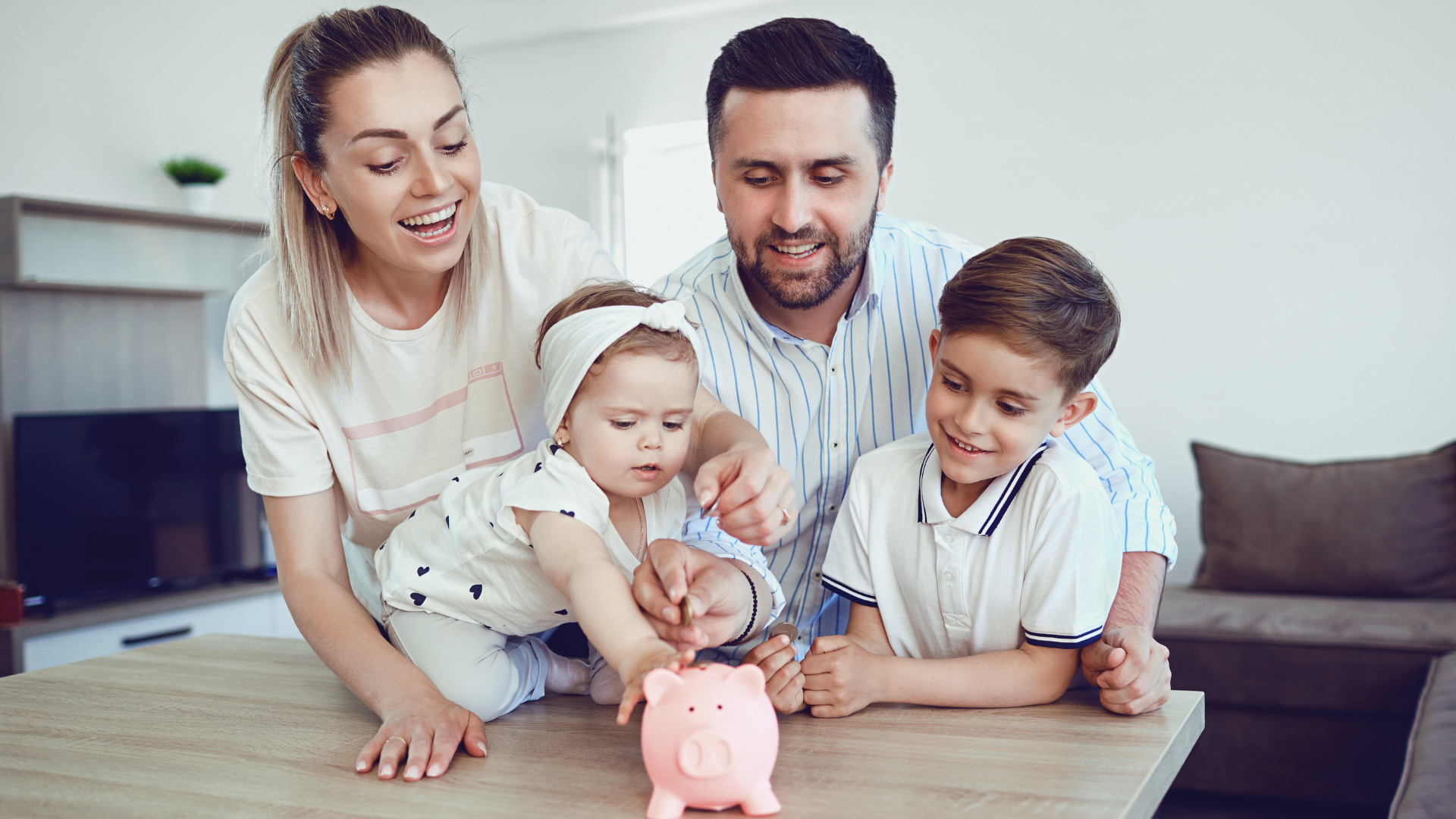 Family of four putting money into a pink piggy bank on a table in a living room.